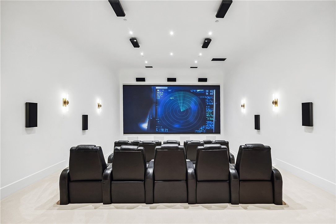 This is an interior shot of a home theater. The room features tiered seating with black leather recliners facing a large screen displaying a radar-like image. The walls are painted white and adorned with sconces and speakers, while the ceiling has recessed lighting and additional speakers, creating a modern and immersive entertainment space.
