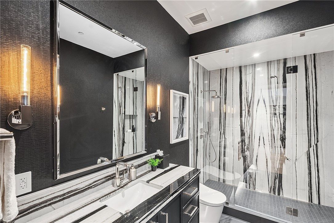 This is a modern primary bathroom featuring a black and white color scheme. The vanity has a marble countertop and dark cabinetry, complemented by a large mirror and stylish sconces. A glass-enclosed shower with striking black and white marble-like tile adds a luxurious touch.