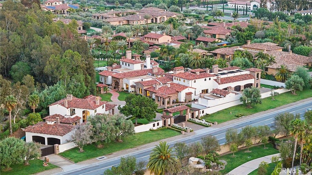 This aerial view showcases a luxurious residential estate with multiple buildings featuring red tile roofs and white stucco walls, surrounded by lush landscaping and mature trees. The property includes manicured lawns, gated entrances, and a winding driveway, creating an impression of privacy and exclusivity. The overall style is reminiscent of Mediterranean architecture.