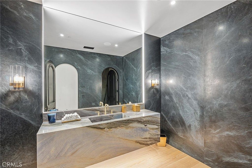 This is a modern bathroom featuring dark marble walls and a unique, multi-toned marble countertop with an integrated sink. A large, illuminated mirror enhances the space, while gold accents add a touch of luxury. The overall design is sleek and sophisticated, creating a spa-like atmosphere.