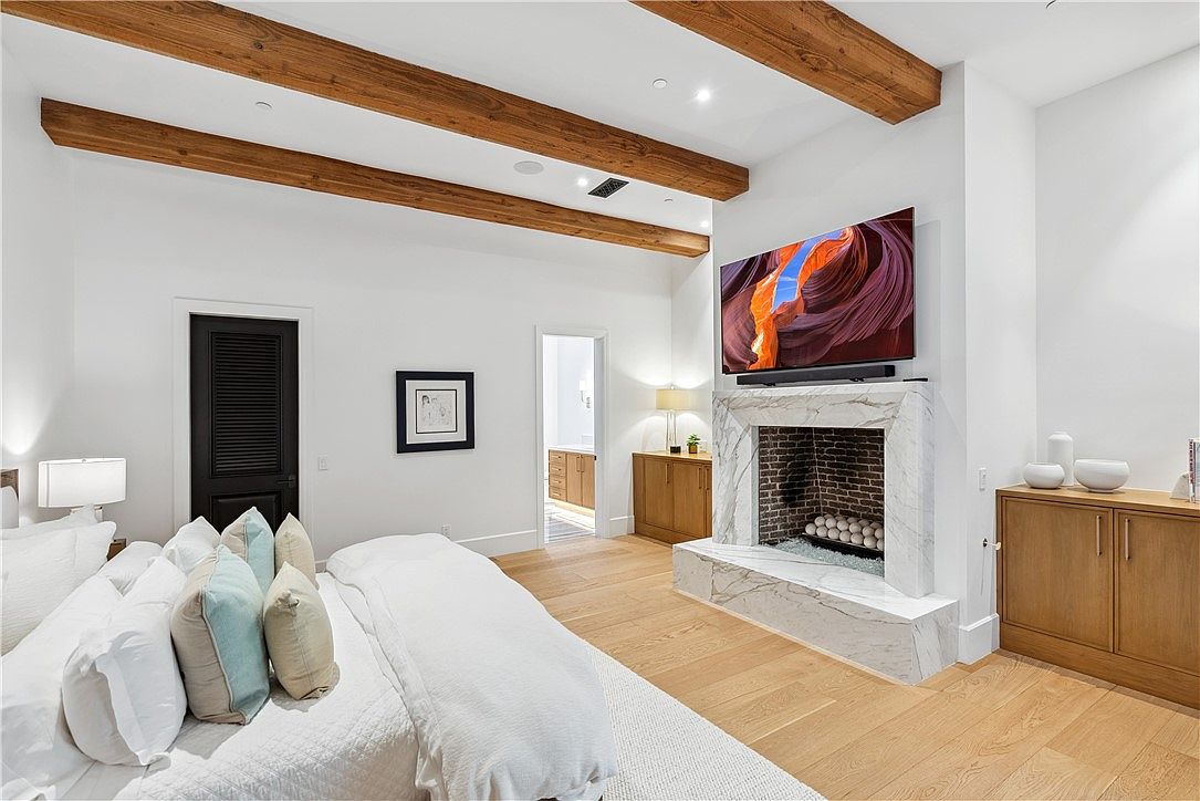 This is a primary bedroom featuring a large bed with white linens and decorative pillows. The room has wooden ceiling beams, a fireplace with a marble surround and a large television mounted above it. There are built-in cabinets on either side of the fireplace and a black louvered door on the left wall.