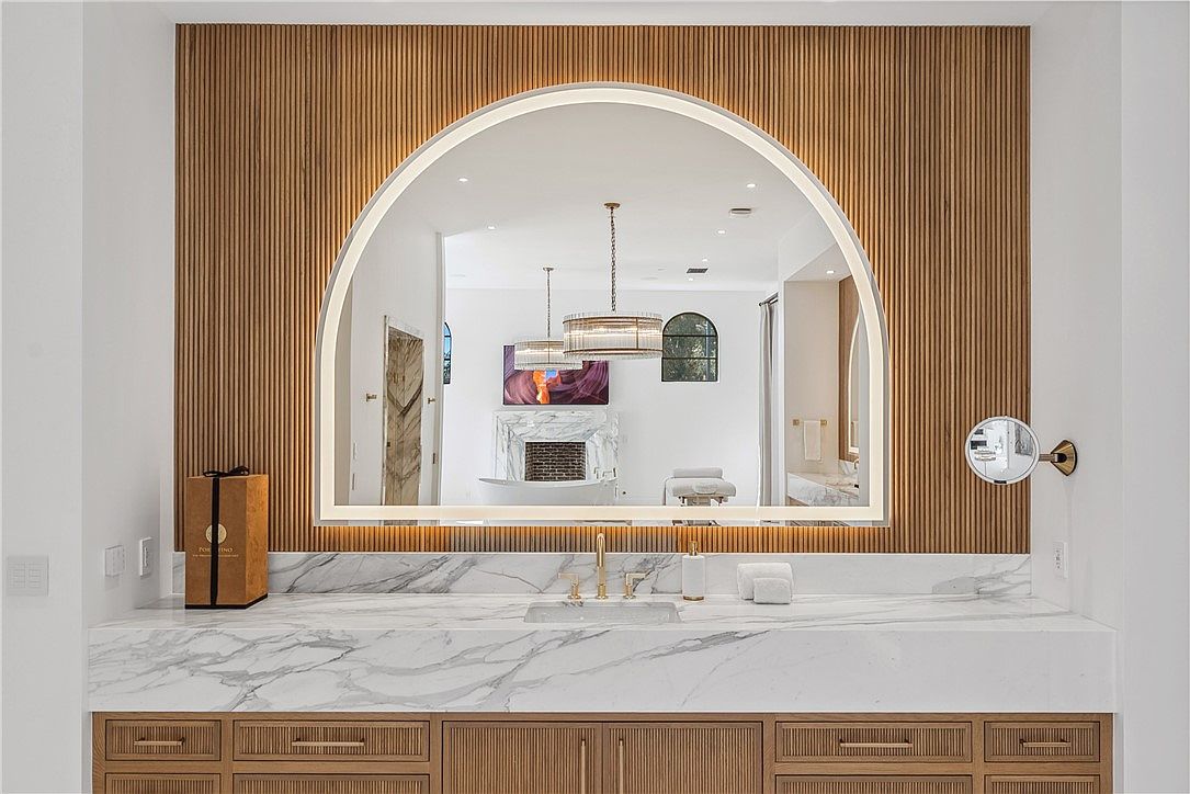 This is a primary bathroom featuring a large, arched mirror with integrated lighting, set against a wood-paneled wall. The vanity has a marble countertop and wood cabinetry with a reeded texture. The mirror reflects a view into the rest of the bathroom, including a fireplace and chandelier, creating a luxurious and spacious feel.