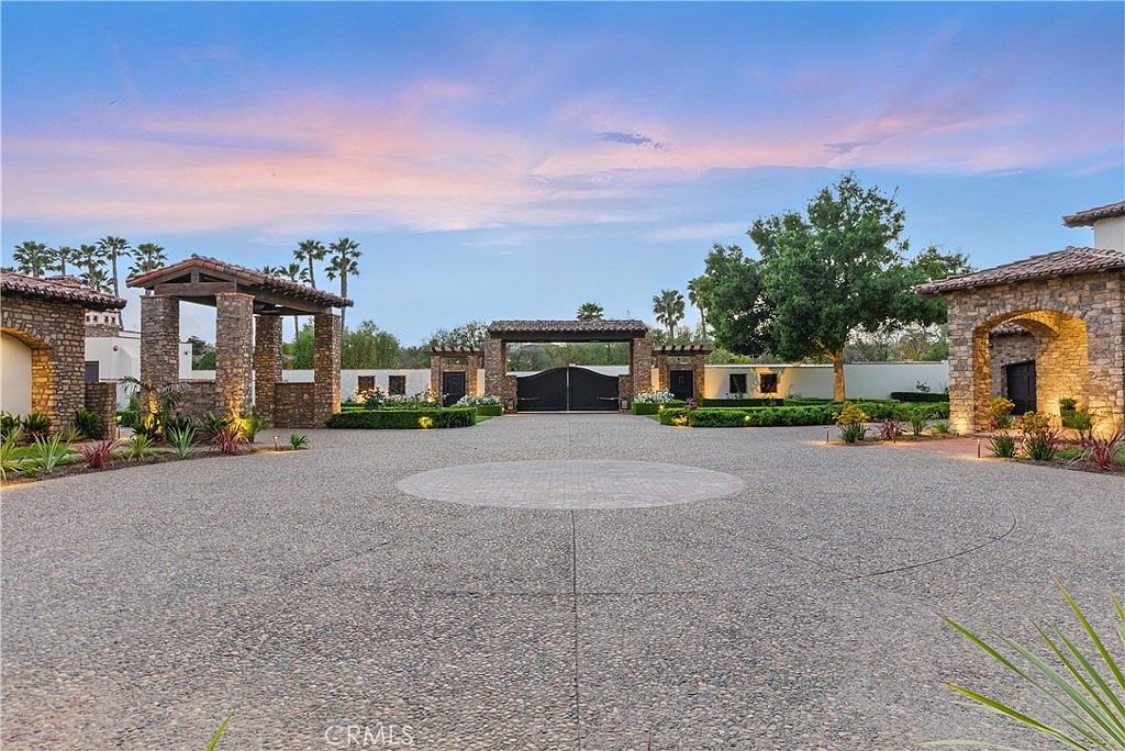 This is a grand front view of a luxurious estate, showcasing a circular driveway made of gravel and brick, leading to an ornate gated entrance. The property features stone archways and meticulously manicured landscaping, creating an impressive and welcoming approach. The sky is a beautiful mix of blues and pinks, suggesting either sunrise or sunset.