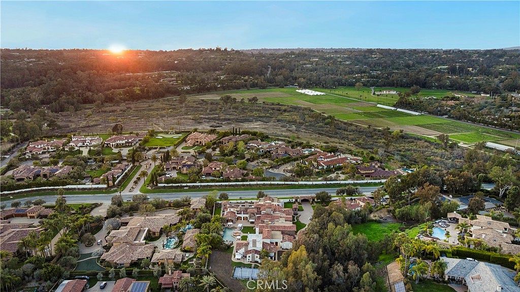 This aerial shot showcases a luxurious residential area with sprawling estates, manicured lawns, and mature trees. The homes feature Mediterranean-style architecture with red tile roofs and lush landscaping. A nearby field adds a touch of rural charm to the upscale neighborhood, creating a serene and exclusive atmosphere.