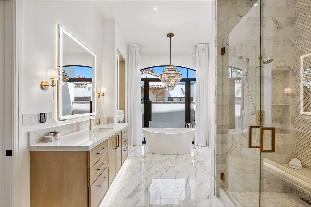 This is a luxurious primary bathroom featuring a double vanity with light wood cabinetry and marble countertops. A freestanding bathtub sits beneath a large arched window with white curtains, and a glass-enclosed shower with marble tile and bronze fixtures is visible to the right. The room is bright and spacious, with marble flooring and a modern chandelier adding to the elegant ambiance.