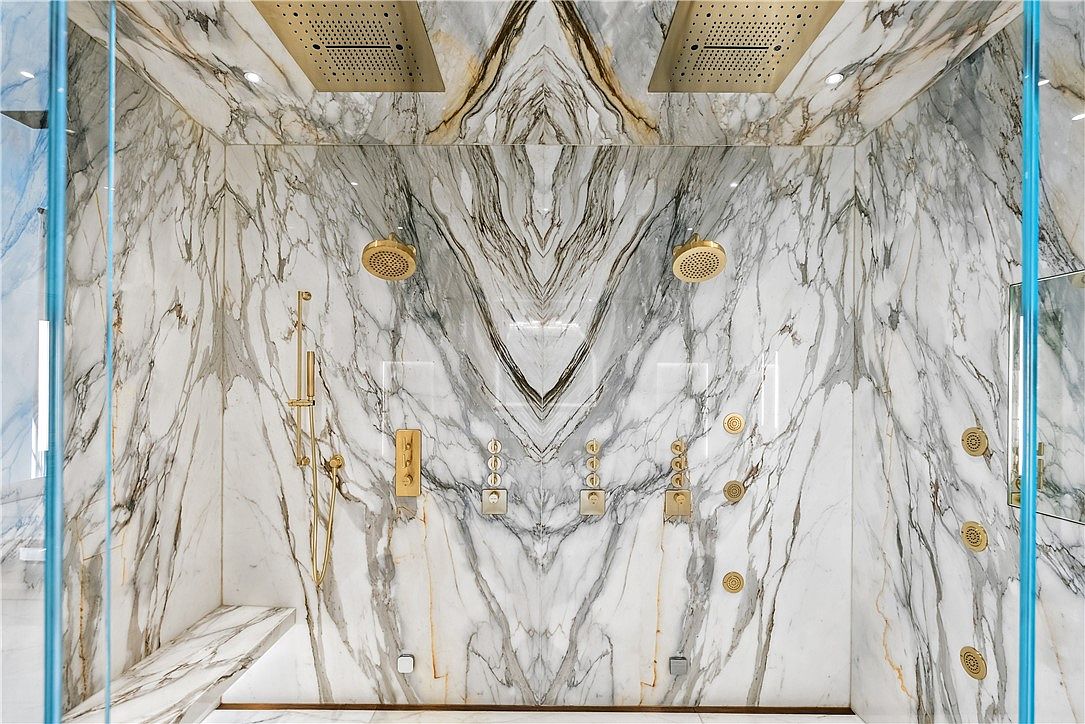 This is a luxurious primary bathroom featuring a spacious walk-in shower with stunning marble walls and gold fixtures. The shower is equipped with multiple shower heads, including overhead rain fixtures and body jets, creating a spa-like experience. Glass enclosures add to the modern and open feel of the space.