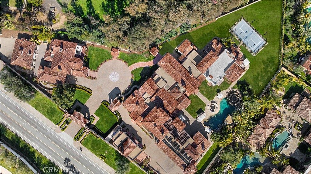This aerial view showcases a sprawling estate with multiple buildings featuring red tile roofs, lush green lawns, and mature trees. A circular driveway leads to the main residence, which includes a pool, spa, and a tennis court. The overall impression is one of luxury and privacy.
