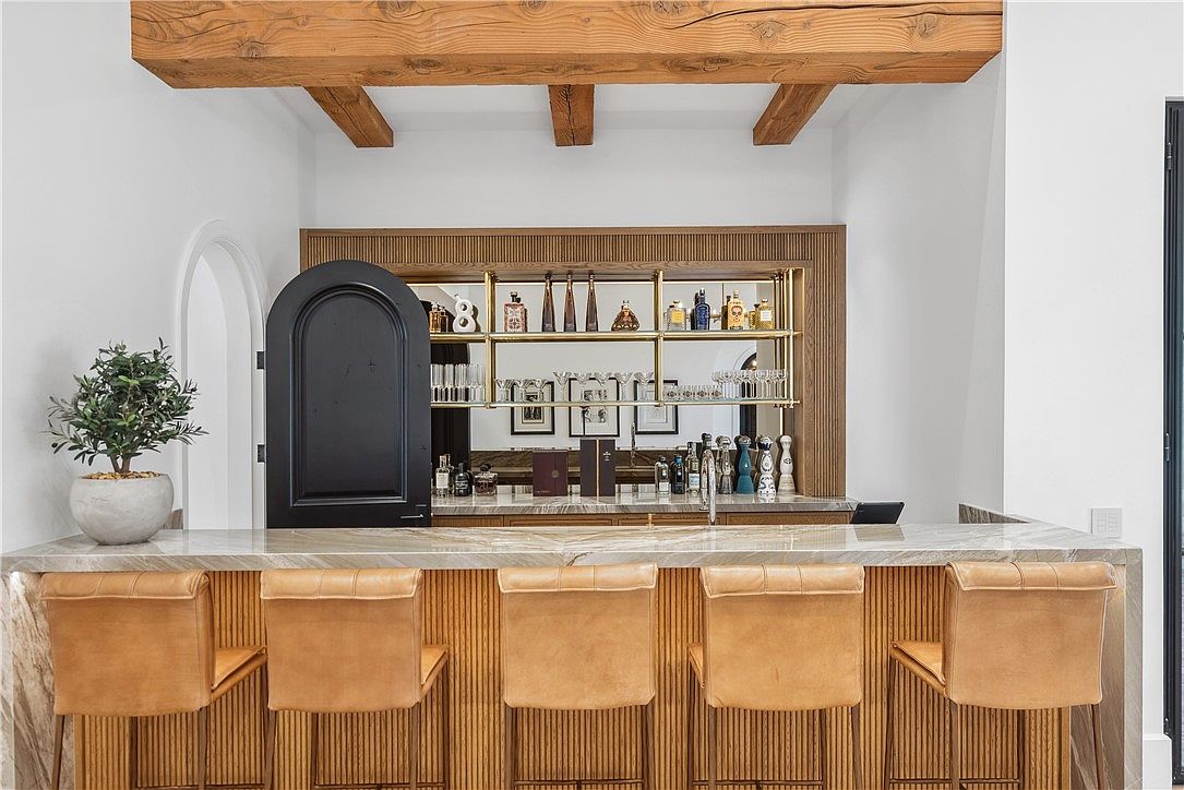 This image showcases a stylish home bar area, featuring a marble countertop with seating for five. The bar is backed by a wooden-framed shelving unit displaying various bottles and glassware, complemented by a black arched door on the left. The design incorporates natural wood elements and a sophisticated color palette, creating an inviting and upscale atmosphere.