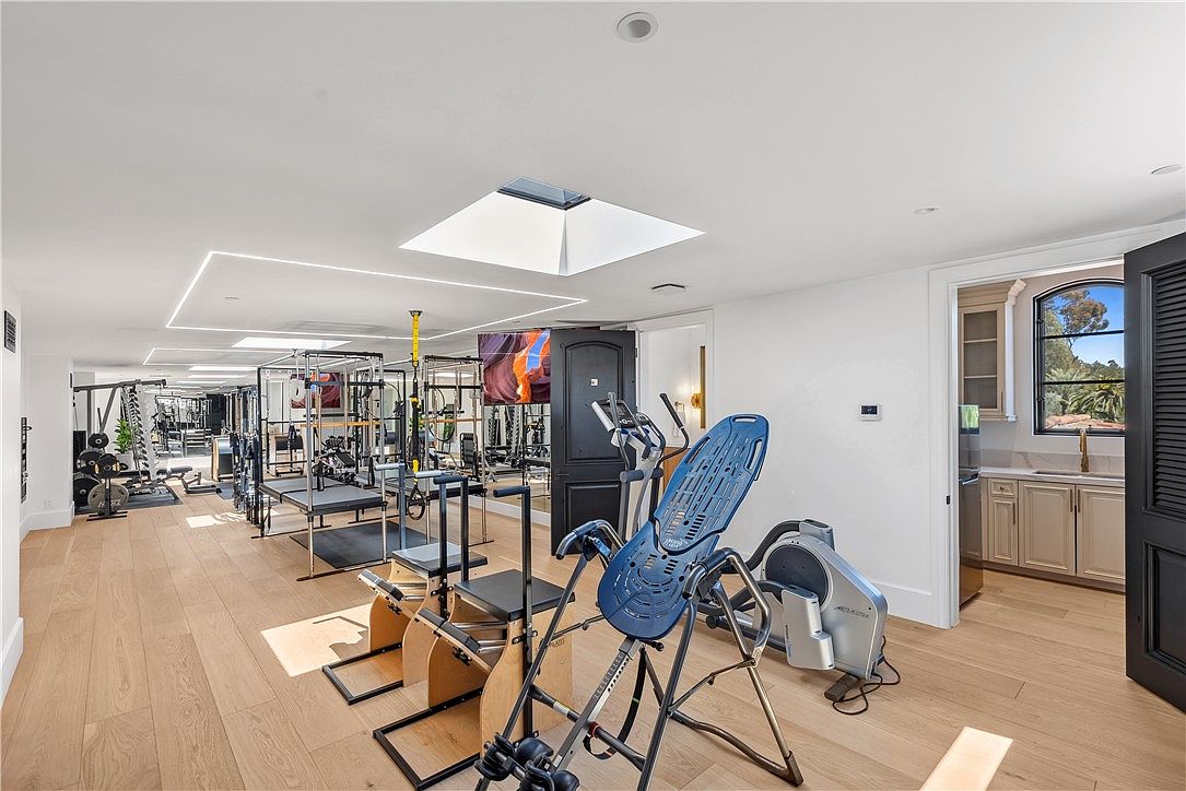 This is a well-equipped home gym featuring a variety of exercise machines and equipment, including weight machines, Pilates reformers, and an inversion table. The room has light wood flooring, white walls, and recessed lighting, with natural light coming from a skylight. The space also includes a television and a doorway leading to a small kitchenette area, creating a functional and luxurious workout environment.