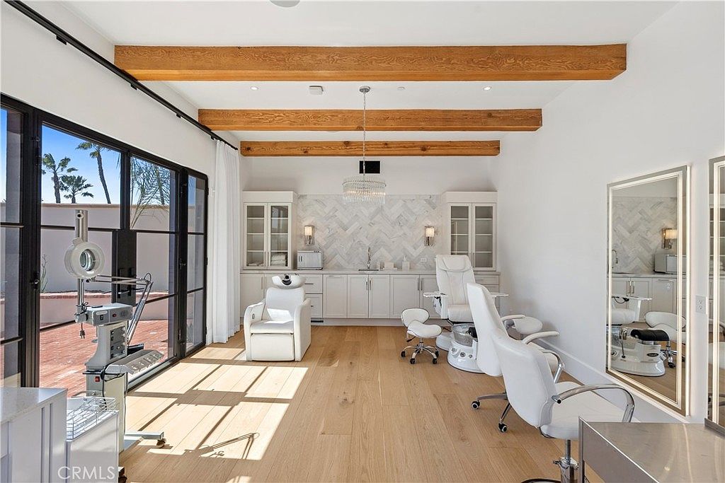 This interior shot showcases a bright and airy salon or spa room, featuring white walls, light wood flooring, and exposed wooden beams on the ceiling. The room is equipped with salon chairs, a sink area with white cabinetry and marble backsplash, and large windows that let in plenty of natural light. The overall impression is clean, modern, and luxurious.