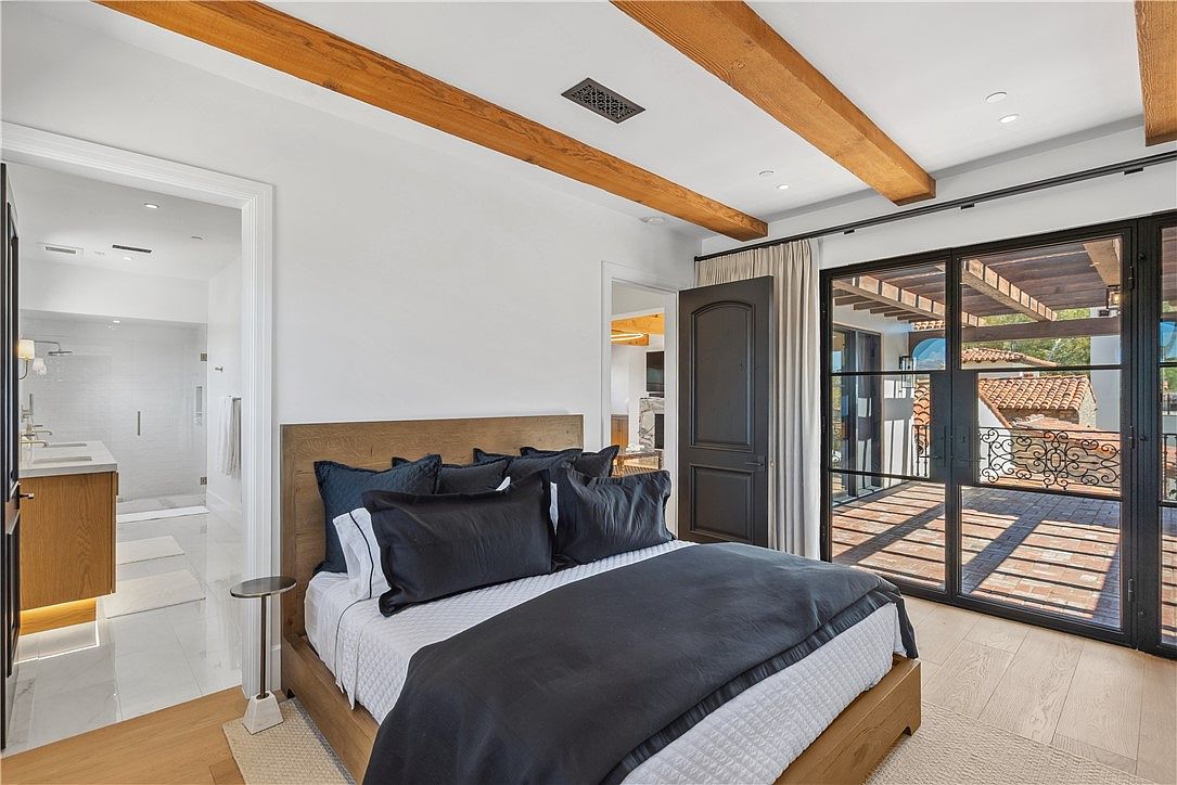 This is a primary bedroom featuring a wooden bed with dark blue pillows and a gray duvet. The room has wooden ceiling beams and a black-framed glass door leading to a balcony. There is also a doorway leading to a bathroom, and the overall style is modern and luxurious.