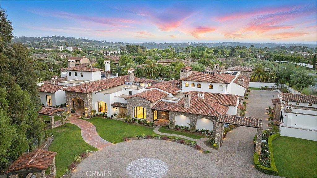 This aerial view showcases a sprawling estate with a terracotta tile roof, white stucco walls, and stone accents. The property features lush landscaping, a circular driveway, and multiple structures, creating a luxurious and inviting atmosphere. The overall impression is one of grandeur and sophisticated design.