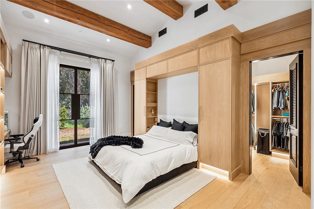 This is a primary bedroom featuring a built-in bed frame with integrated storage and lighting. The room has a neutral color palette with wooden beams on the ceiling and a large window providing natural light. A walk-in closet is accessible from the bedroom, adding to the convenience and luxury of the space.