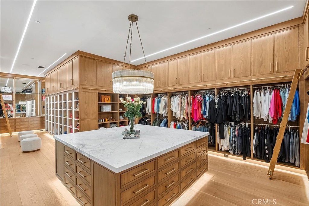This is a luxurious walk-in closet featuring custom wood cabinetry and drawers, providing ample storage space for clothing and accessories. A large marble-topped island sits in the center, topped with a vase of flowers, while a decorative chandelier hangs above. The closet is well-lit with recessed lighting and under-cabinet lighting, creating a bright and inviting space.
