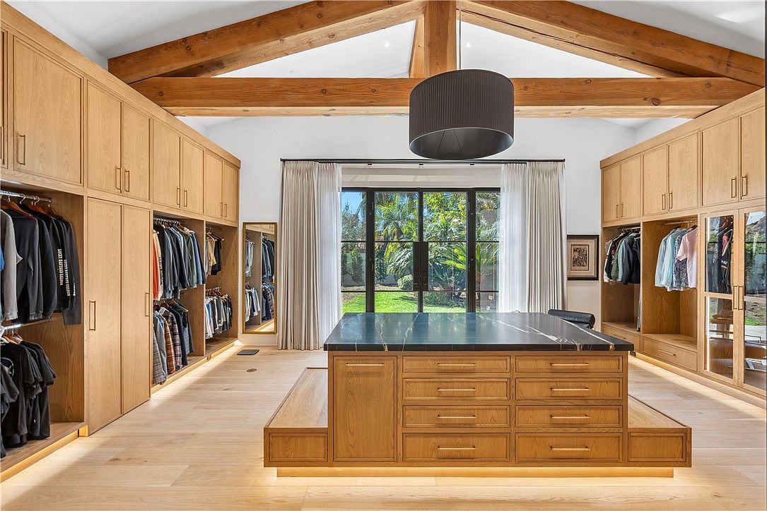This is a spacious walk-in closet featuring custom wood cabinetry and a large center island with a dark countertop. The closet is well-organized with hanging clothes and drawers, and the design incorporates natural light through a large window. The overall impression is luxurious and functional, perfect for a high-end real estate listing.
