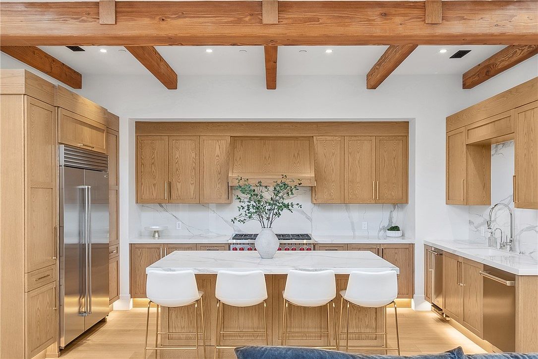 This is a bright and modern kitchen featuring light wood cabinetry, stainless steel appliances, and a large island with a white countertop and seating for four. The kitchen is well-lit with recessed lighting and natural light, and the white marble backsplash adds a touch of elegance. The overall impression is clean, spacious, and inviting.
