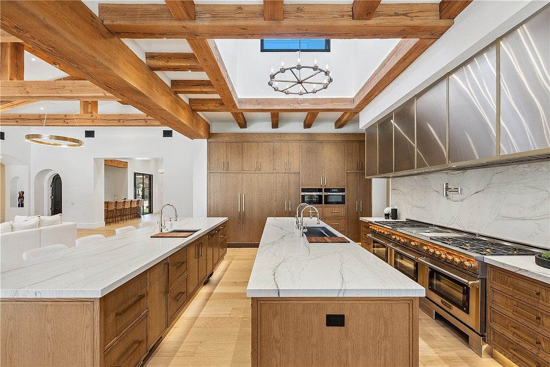 This is a spacious, high-end kitchen featuring two large islands with white marble countertops and wooden cabinetry. The kitchen boasts professional-grade stainless steel appliances, including a large range with a custom hood. Exposed wooden beams adorn the ceiling, adding a rustic touch to the modern design.