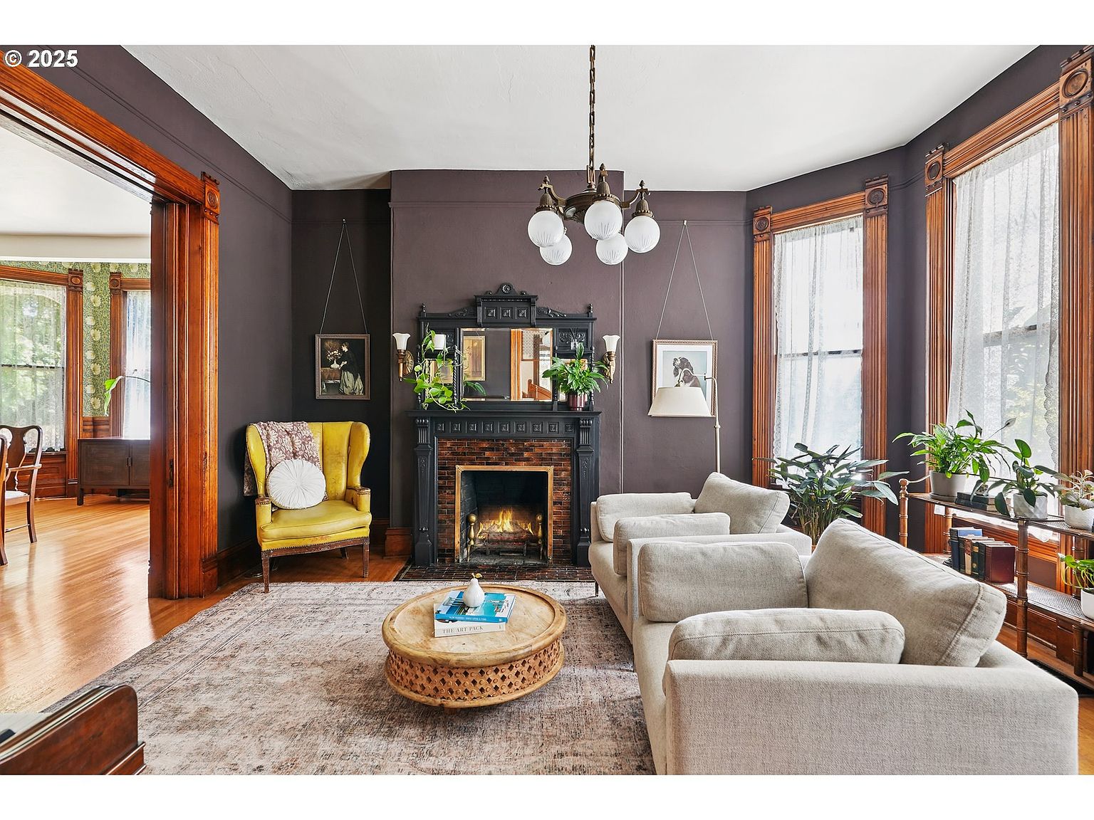 This is an interior shot of a living room featuring dark brown walls, hardwood floors, and a patterned area rug. The room is furnished with a light-colored sofa, a yellow armchair, and a round coffee table. A fireplace with a decorative mantel serves as a focal point, and natural light streams in through large windows, creating a warm and inviting atmosphere.