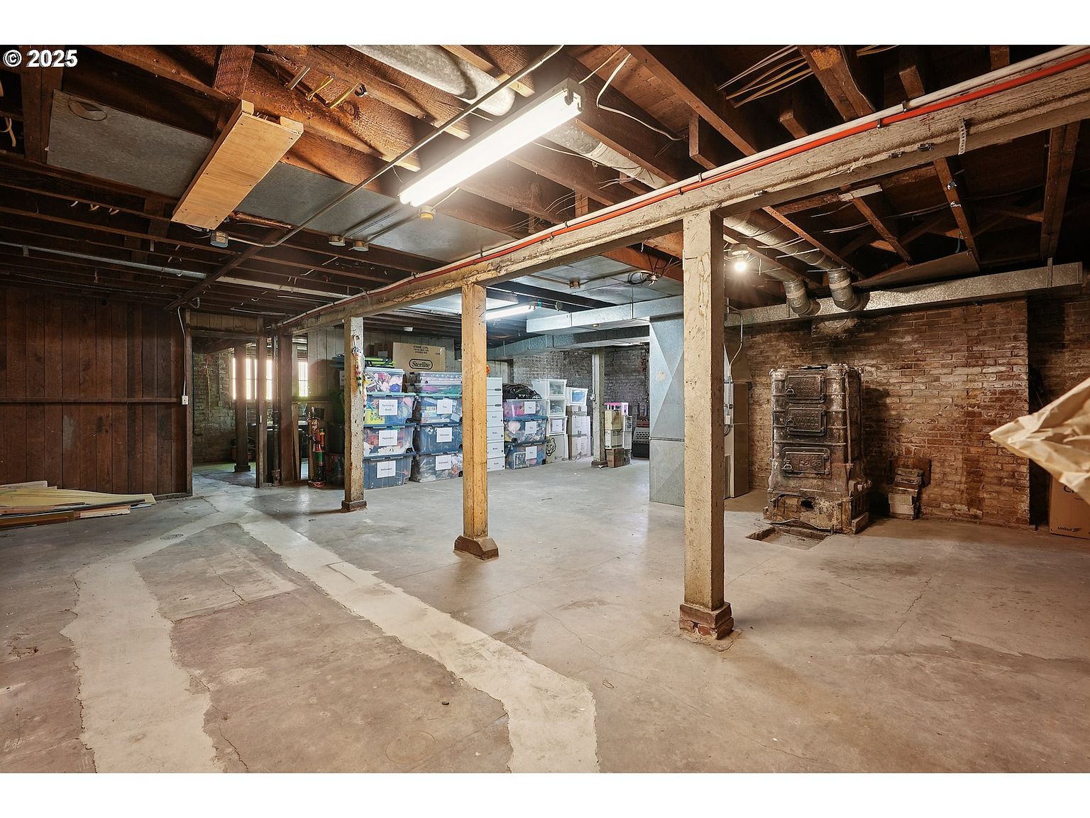 This is a spacious basement featuring exposed wooden beams and support columns. The concrete floor shows signs of wear, and the brick wall adds a rustic touch. Storage boxes are neatly stacked, and an old furnace stands against the brick wall, creating a unique focal point.