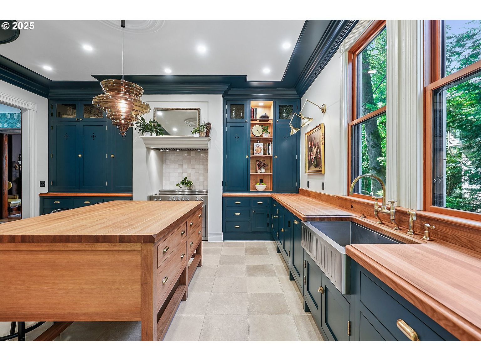 This is a well-lit kitchen featuring custom blue cabinetry with brass hardware and butcher block countertops. A large wooden island stands in the center of the room, and a stainless steel farmhouse sink is installed beneath a window that provides natural light. The kitchen also includes a decorative range hood and a unique, multi-tiered chandelier.