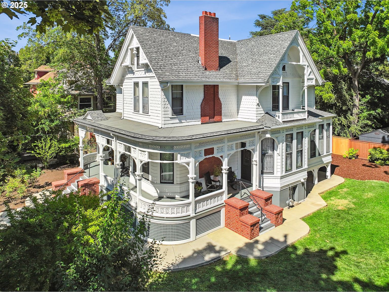 This is an eye-level aerial shot of a charming two-story Victorian house with a gray roof and white exterior. The house features a prominent red brick chimney, a wraparound porch with intricate detailing, and well-maintained landscaping with green grass and mature trees. The overall impression is one of classic elegance and curb appeal.