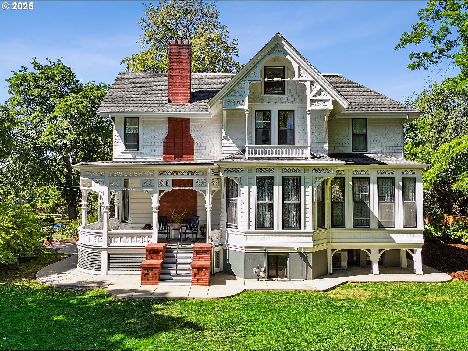 This is a front exterior view of a large, ornate Victorian-style house. The house features a prominent red brick chimney, a wraparound porch with decorative woodwork, and multiple windows with dark frames. The well-manicured lawn and mature trees add to the property's curb appeal, creating an inviting and stately impression.