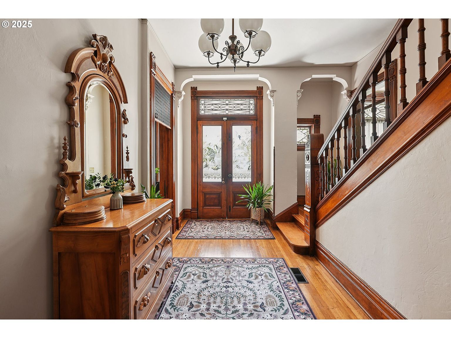 This interior shot showcases a grand hallway with a beautifully crafted wooden staircase and ornate details. A large antique mirror and dresser adorn one side, while a decorative rug leads to a wooden front door with frosted glass. The space exudes a sense of classic elegance and historical charm.