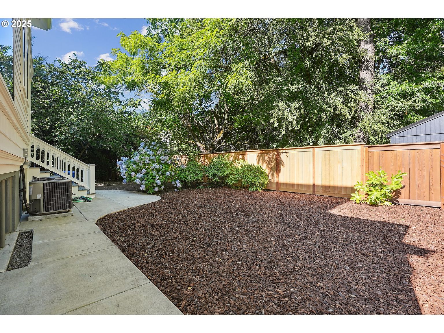 This image showcases a well-maintained backyard featuring a lush garden area with wood chip ground cover, a newly constructed wooden fence, and mature trees providing ample shade. A concrete patio extends from the side of the house, offering a transition from indoor to outdoor living. The yard appears private and inviting, perfect for relaxation and outdoor activities.