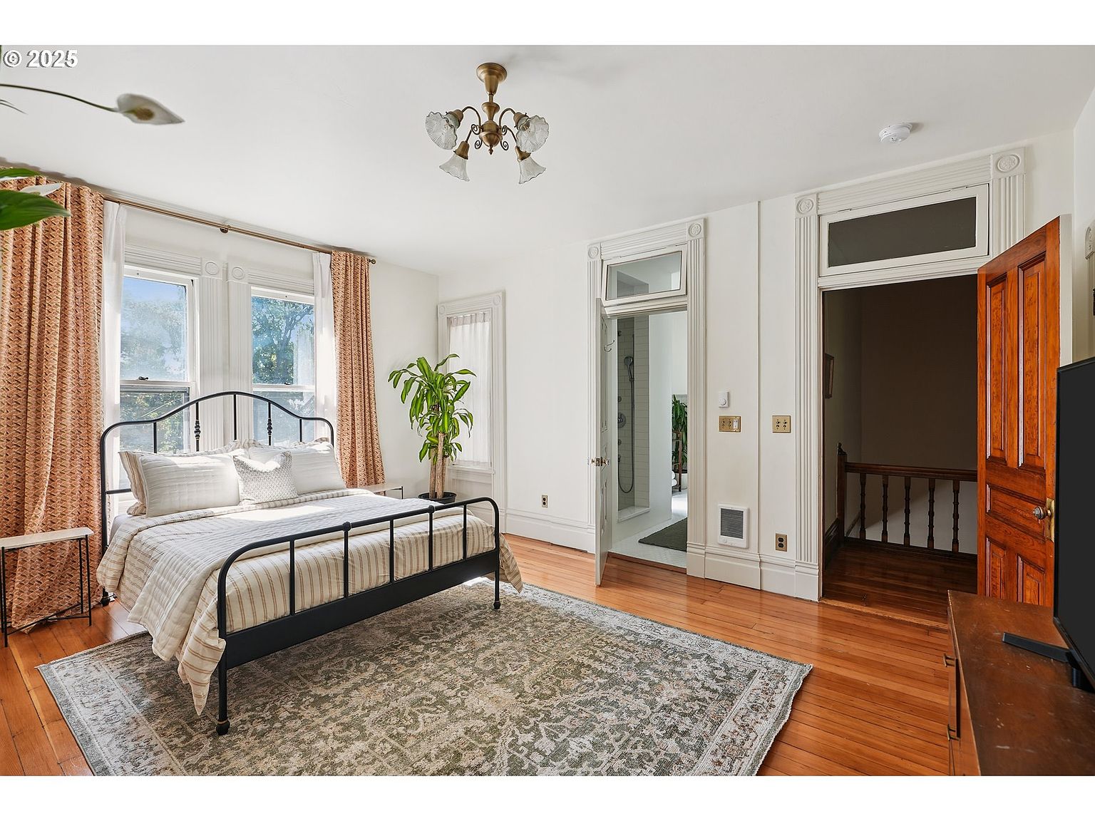 This is a primary bedroom featuring a black metal bed frame with white linens and pillows, set upon a patterned area rug on hardwood floors. Natural light streams in through the windows, complemented by a decorative chandelier. The room also includes access to a bathroom and a hallway/stairwell, showcasing the home's layout and architectural details.