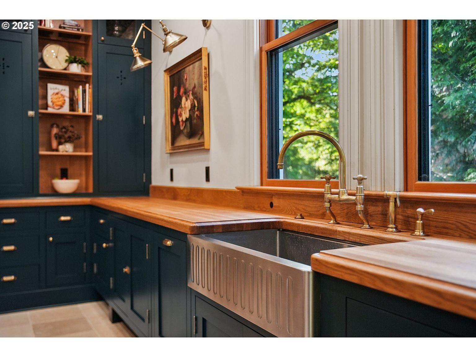 This is a detailed interior shot of a kitchen, showcasing dark blue cabinetry with wooden countertops and brass hardware. A stainless steel farmhouse sink is prominently featured beneath a window that offers a view of lush greenery. The overall aesthetic is a blend of classic and modern, creating a warm and inviting atmosphere.