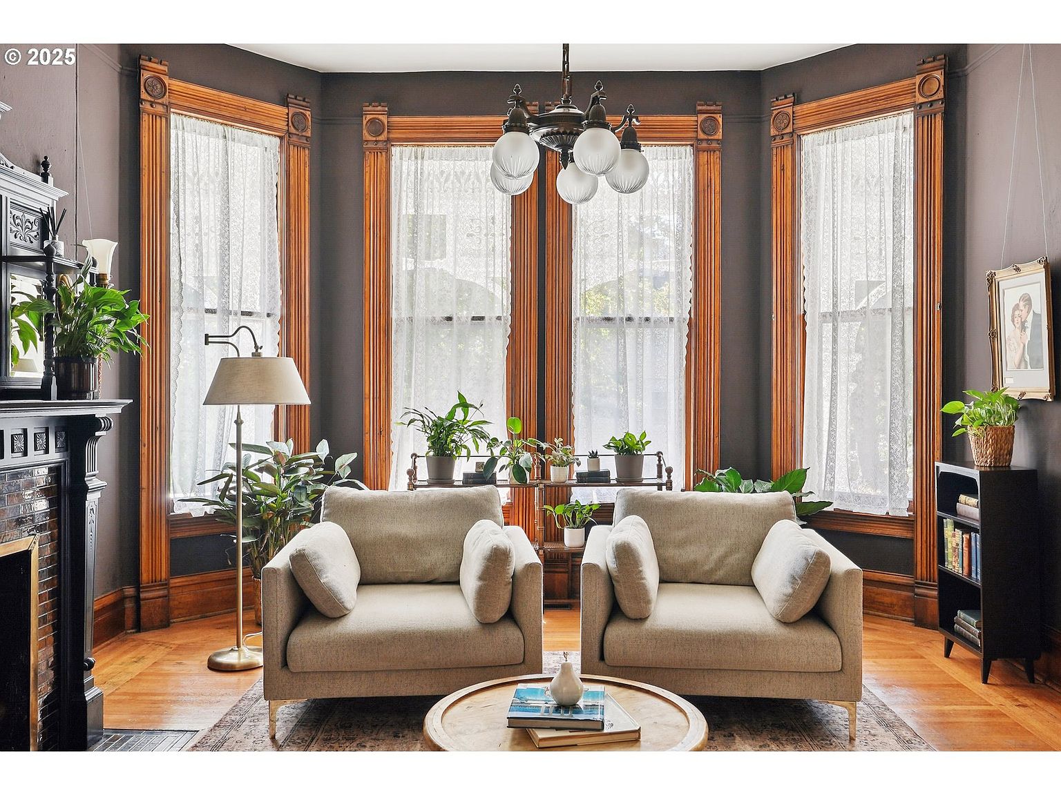 This is an interior shot of a living room featuring two armchairs, a round coffee table, and large windows with lace curtains. The room has a vintage aesthetic with dark walls, wooden trim, and a decorative fireplace. Plants are placed throughout the room, adding a touch of nature to the space.