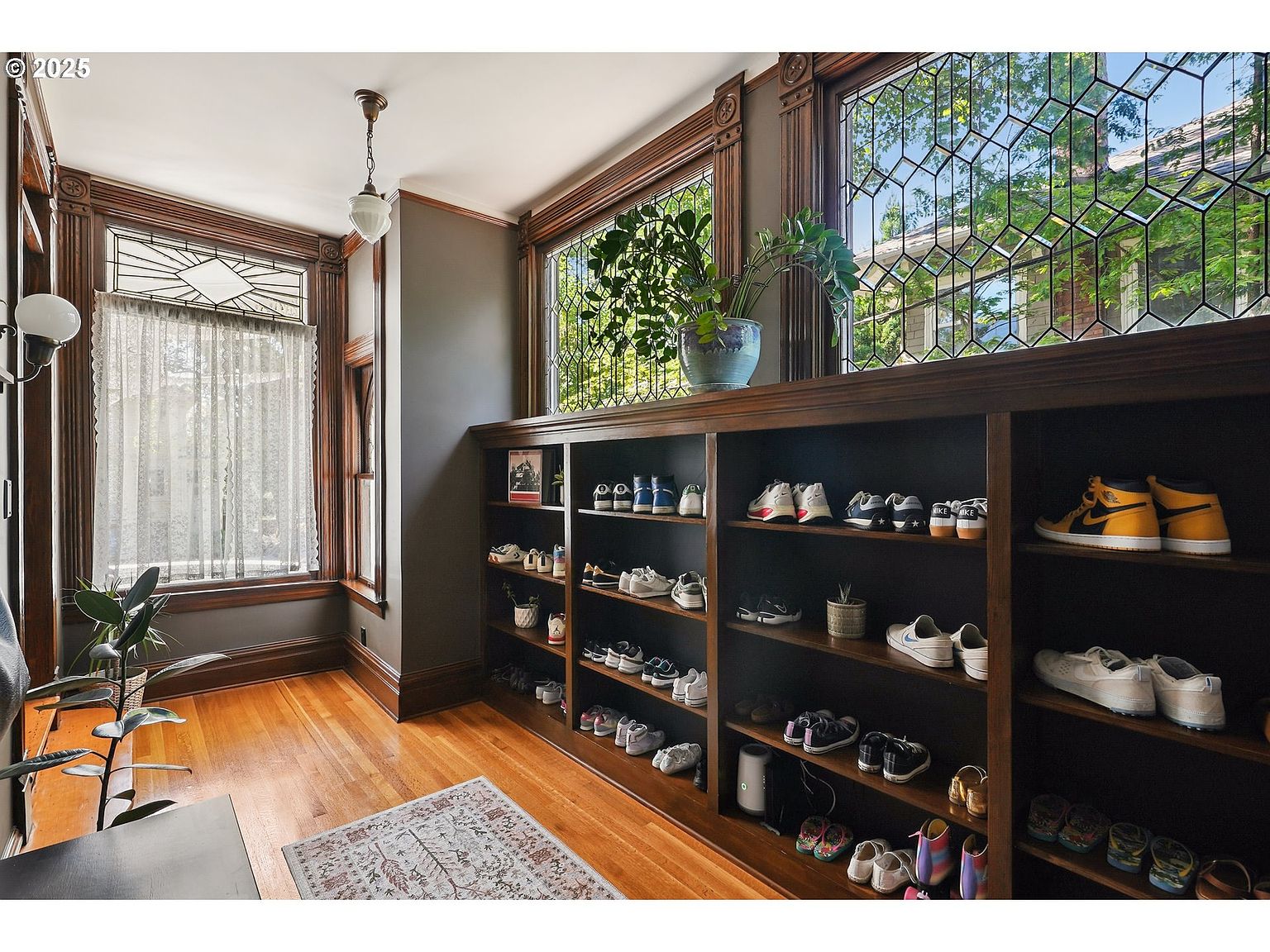 This interior shot showcases a hallway area with rich, dark wood trim and hardwood floors, creating a warm and inviting atmosphere. A custom built-in shelving unit displays an array of shoes, adding a unique and functional element to the space. Large, decorative windows provide ample natural light, enhancing the room's character and charm.