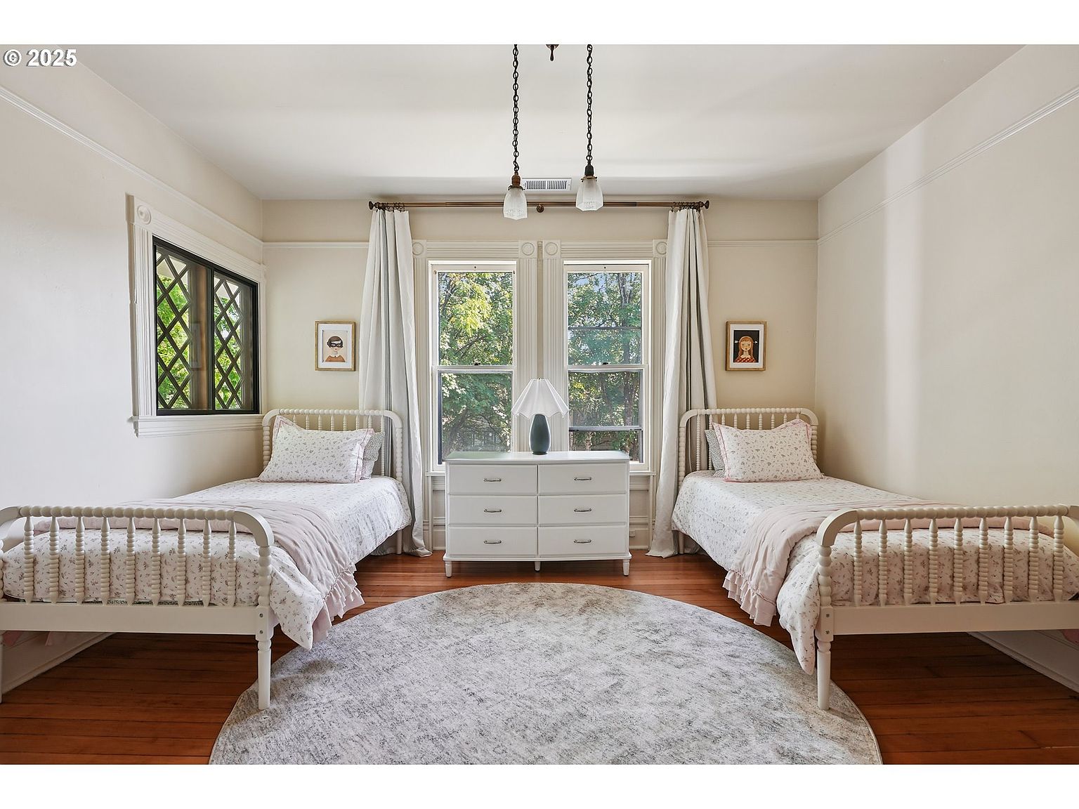This is a charming guest bedroom featuring two twin beds with white frames and floral bedding. A white dresser sits between two windows adorned with light-colored curtains. The room is bright and airy, with hardwood floors and a round area rug adding warmth and character.