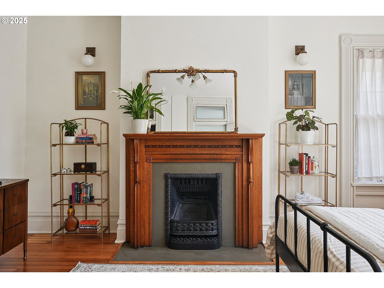 This interior shot showcases a bedroom with a prominent fireplace featuring a wooden mantel and a decorative mirror above. The room is decorated with a blend of classic and cozy elements, including shelving units on either side of the fireplace displaying books and plants. The overall impression is one of warmth and refined comfort, suitable for a primary bedroom.