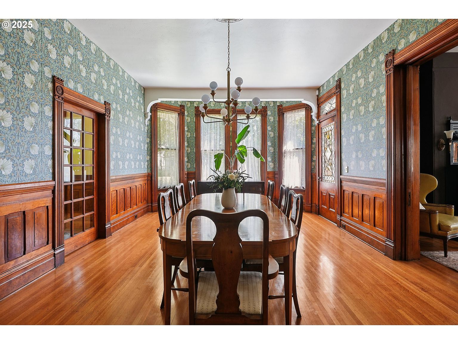 This is a dining room featuring hardwood floors, wood paneling, and floral wallpaper. A large wooden dining table with chairs is centered in the room, illuminated by a chandelier. The room has a classic, elegant style with detailed architectural features.