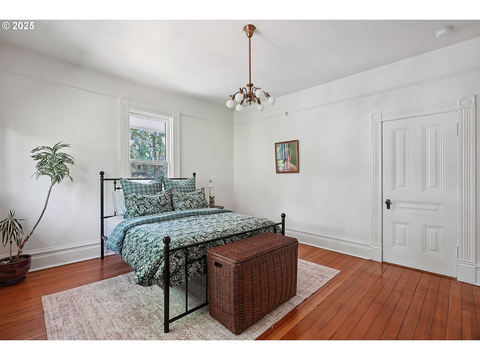 This is a cozy bedroom featuring a black metal bed frame with a green and blue floral patterned comforter and matching pillows. A woven storage chest sits at the foot of the bed, and a patterned rug covers the hardwood floors. The room is painted white with decorative molding, and a window provides natural light, creating a bright and inviting space.