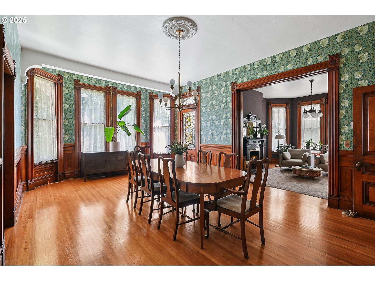 This is an interior shot of a formal dining room featuring hardwood floors, wood-paneled walls with patterned wallpaper above, and a large wooden dining table with chairs. A sideboard sits against the wall beneath a set of windows dressed with sheer curtains, and an open doorway leads to a living room with a fireplace and seating area. The room exudes a sense of classic elegance and spaciousness.