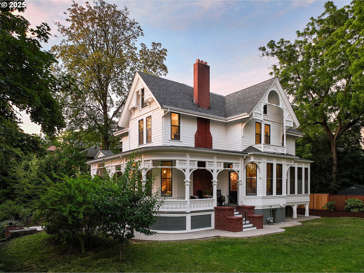 This is a charming front view of a large, white Victorian-style house. The house features a prominent red brick chimney, a wraparound porch with intricate detailing, and multiple gabled roofs. Lush green lawn and mature trees surround the property, adding to its curb appeal and creating a sense of established elegance.