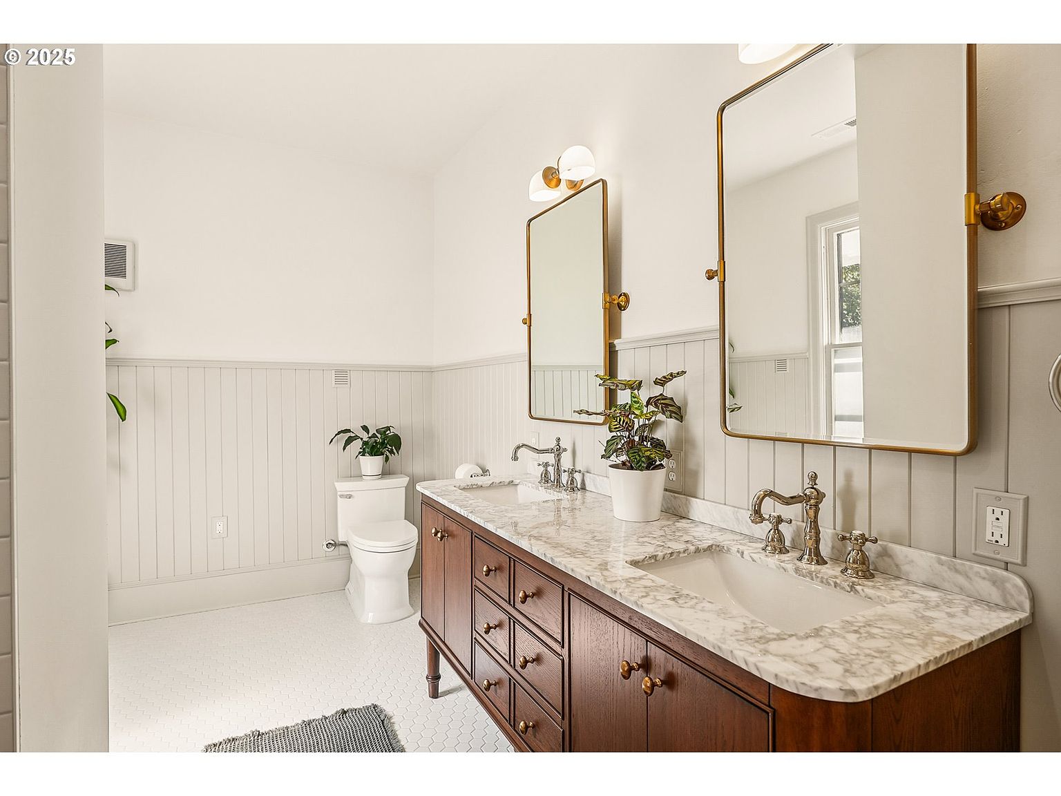 This is a well-lit primary bathroom featuring a double vanity with a dark wood cabinet and marble countertop. Two rectangular mirrors with gold frames hang above the sinks, complemented by sconce lighting. The walls are paneled with white wainscoting, and the floor is tiled with small white hexagonal tiles, creating a clean and classic aesthetic.