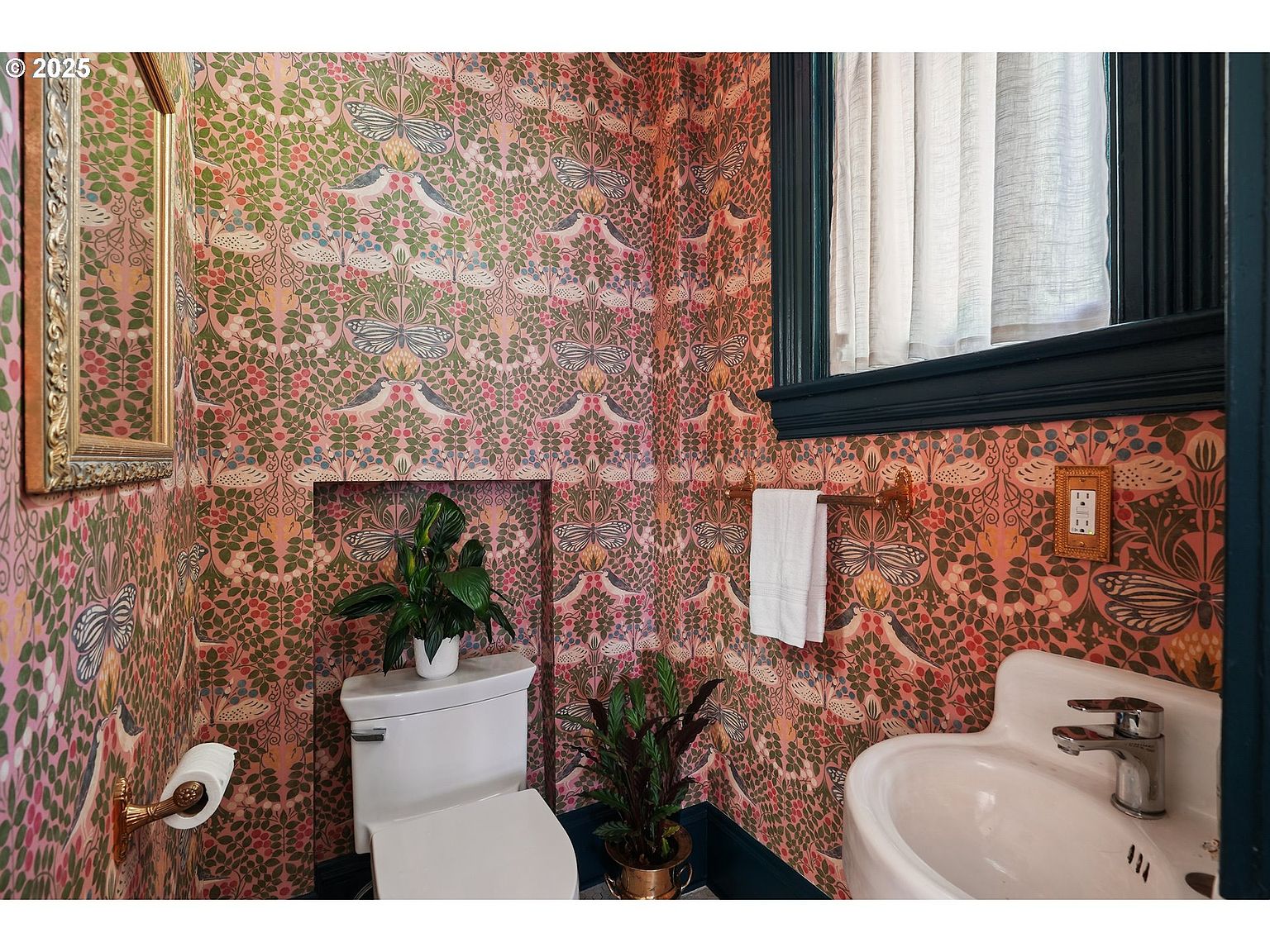 This is a charming guest bathroom featuring a unique, patterned wallpaper with birds and floral motifs. The room includes a toilet, a pedestal sink, and a framed mirror, all complemented by dark blue trim around the window and baseboards. The overall impression is a cozy and stylish space with a vintage touch.
