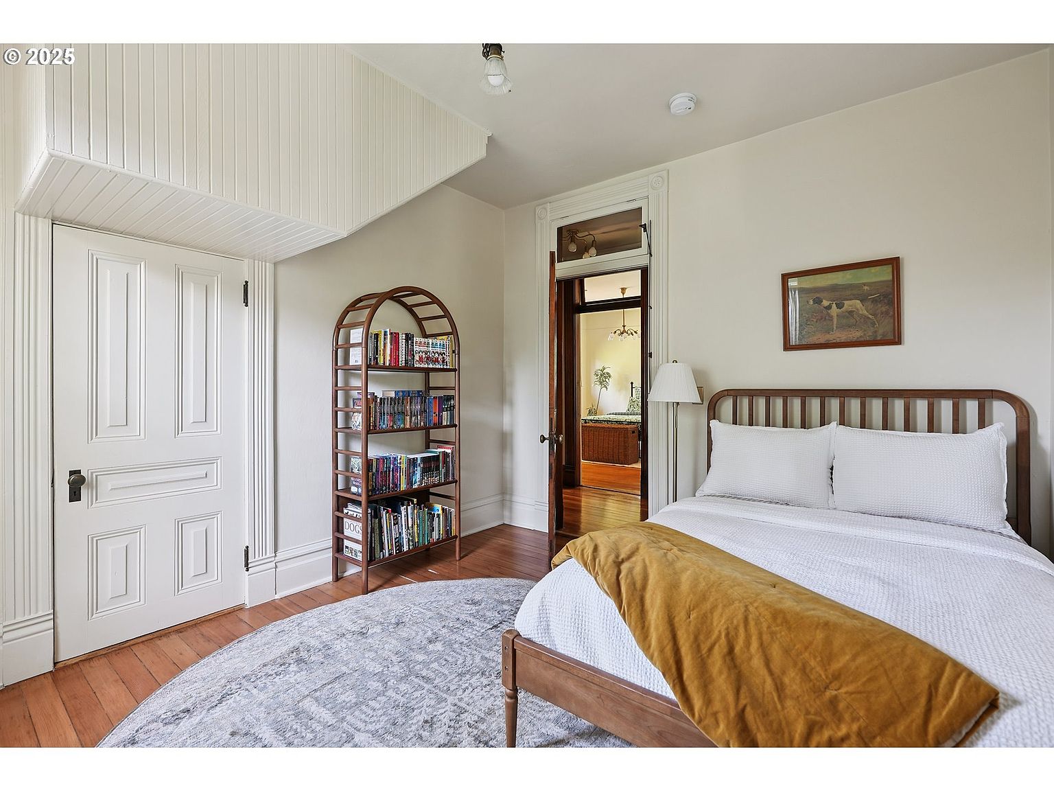 This is a cozy bedroom featuring a wooden-framed bed with white linens and a mustard-colored throw blanket. A round rug sits on the hardwood floor, and a bookshelf filled with books adds character to the room. The walls are painted in a neutral tone, and natural light filters in, creating a warm and inviting atmosphere.