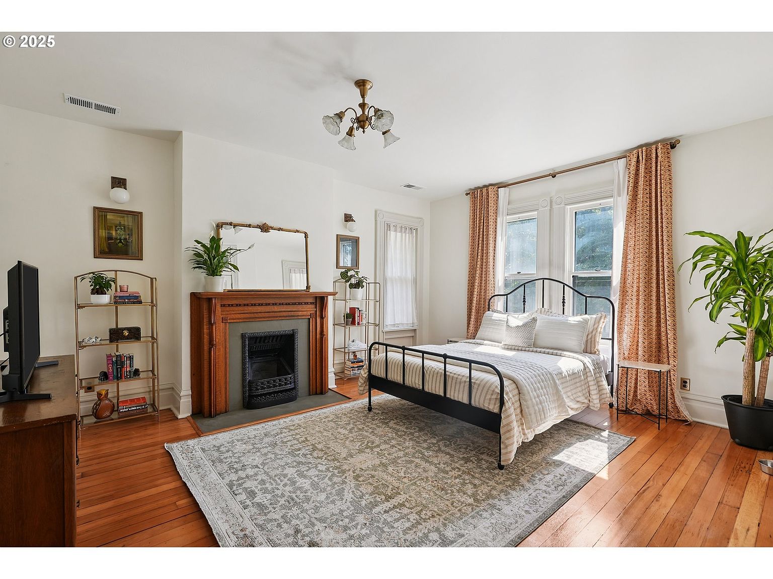 This is a bright and airy primary bedroom featuring hardwood floors, a decorative fireplace, and a wrought iron bed frame. The room is decorated in neutral tones with patterned curtains and a large area rug, creating a cozy and inviting atmosphere. Natural light floods the space through the windows, enhancing the room's appeal.