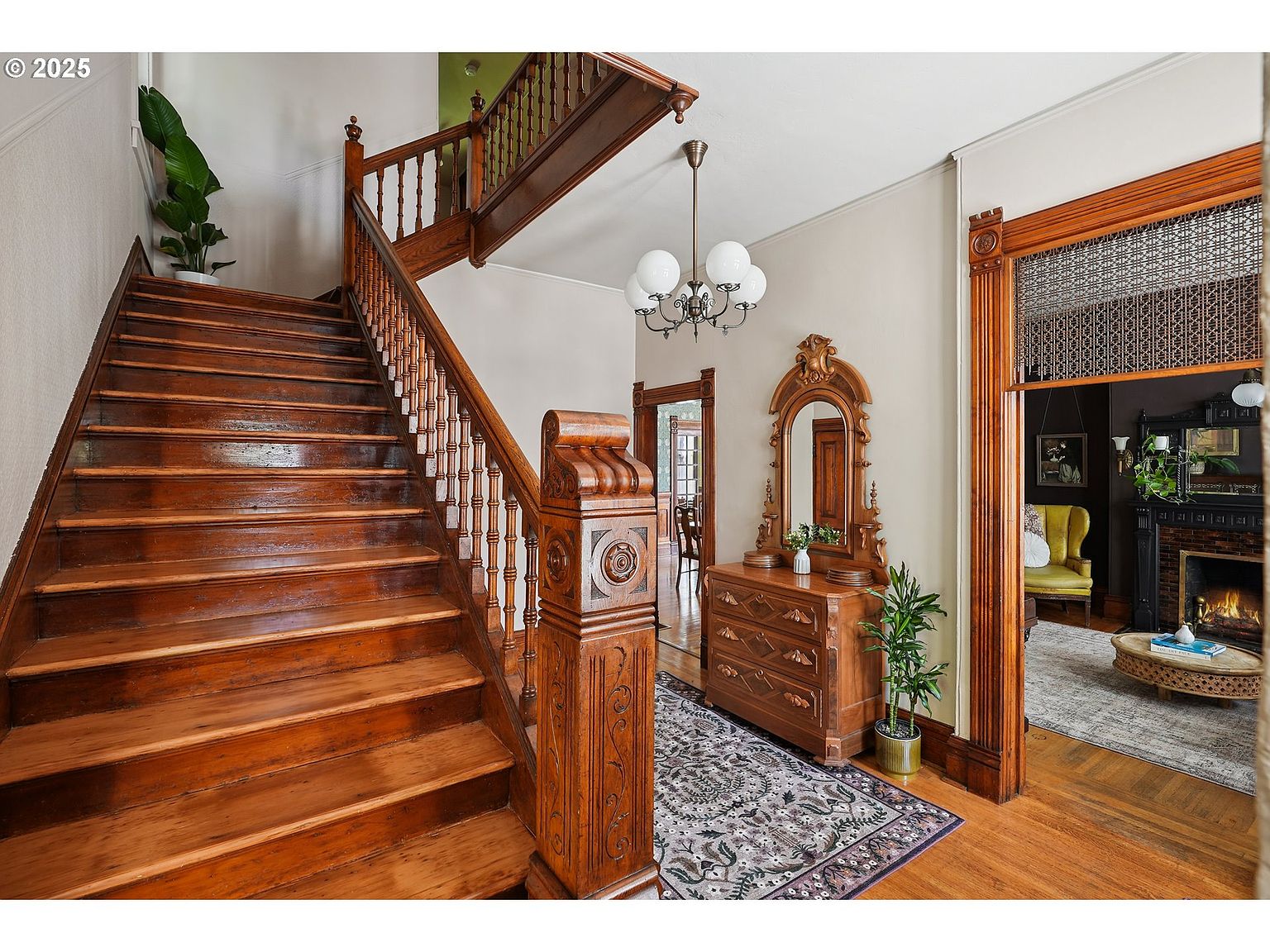 This interior shot showcases a grand hallway with a prominent wooden staircase, featuring detailed carvings and a rich, warm tone. A vintage-style dresser with a mirror sits against the wall, complemented by a patterned rug and a view into a cozy living room with a fireplace. The overall impression is one of classic elegance and historical charm.