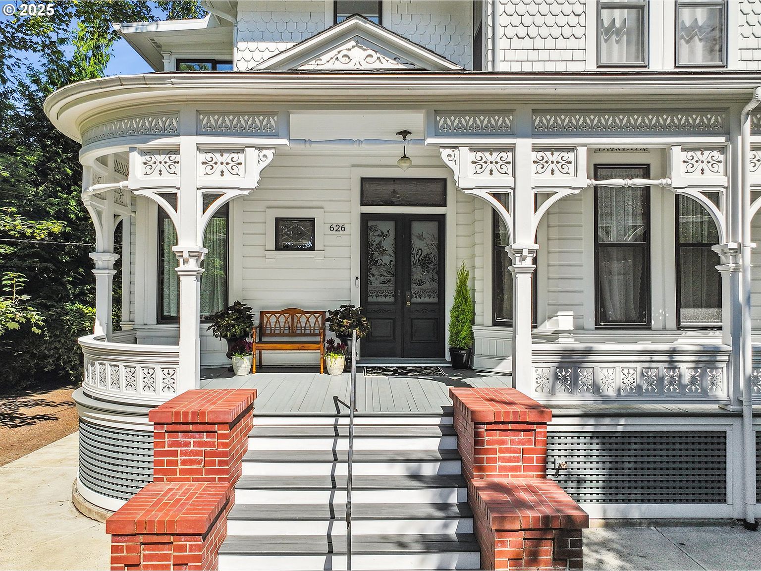This is a welcoming front entryway of a charming Victorian-style home. The entrance features a detailed porch with ornate white columns and decorative trim, leading to a dark double door with glass panels. Brick steps with a metal handrail guide visitors to the porch, and potted plants add a touch of greenery to the scene.