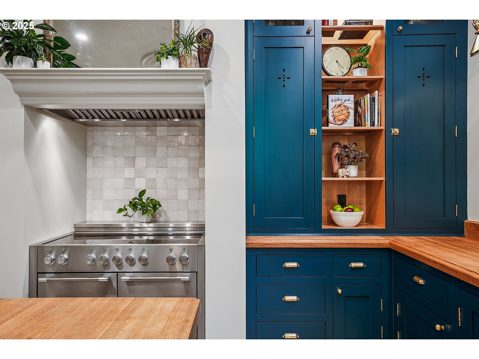 This image showcases a well-appointed kitchen featuring a professional-grade stainless steel range with a custom hood and tiled backsplash. Adjacent to the cooking area is a striking built-in cabinet in a deep blue hue, complemented by wooden countertops and open shelving displaying decorative items and cookbooks. The overall design exudes a blend of classic and contemporary styles, suggesting a high-end and functional culinary space.