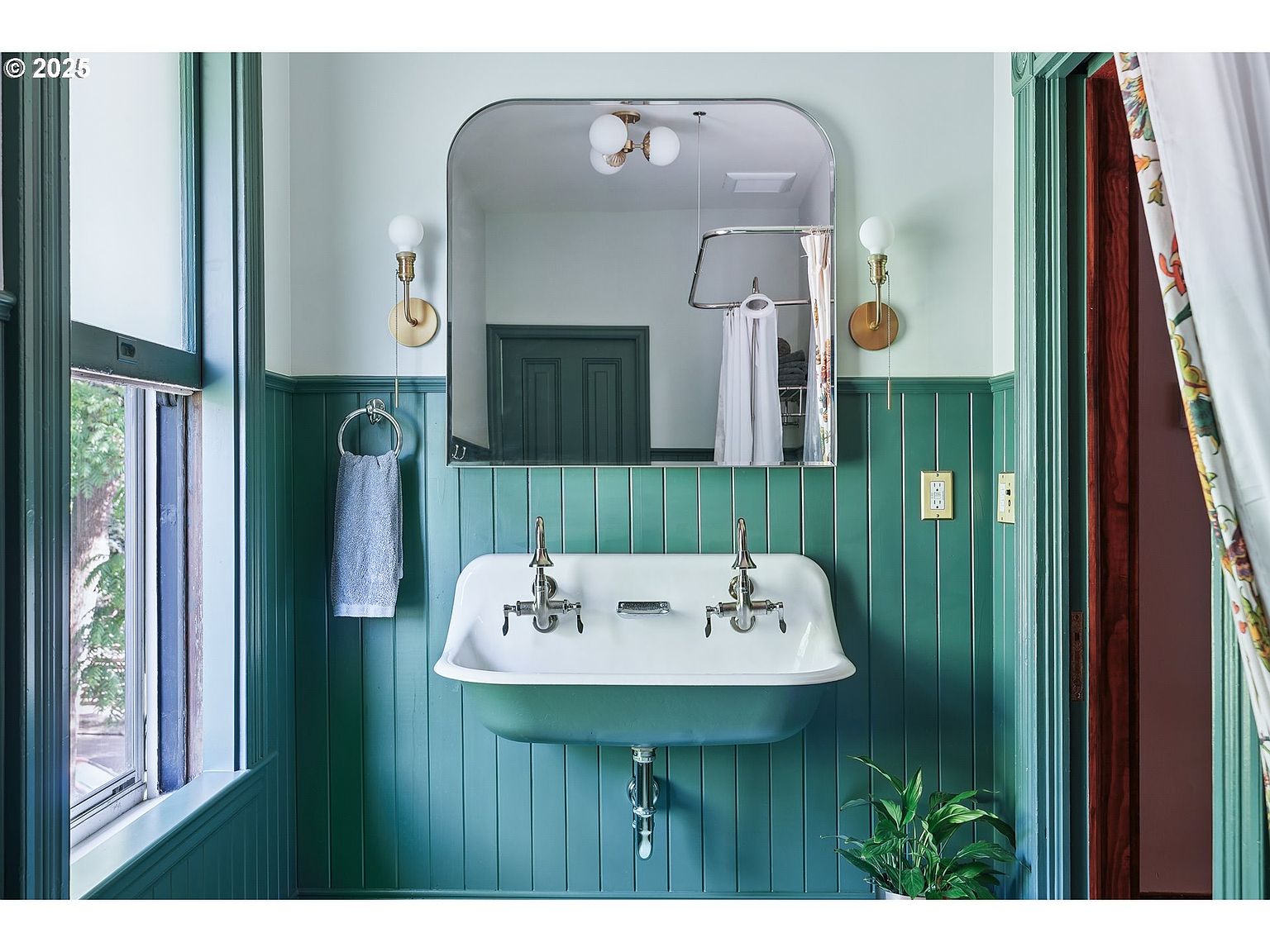 This is a charming bathroom featuring a vintage-style sink with dual faucets, set against a backdrop of teal-painted vertical paneling. A large, arched mirror reflects the room, enhancing the sense of space. The room is well-lit by sconces on either side of the mirror, and a plant adds a touch of nature.