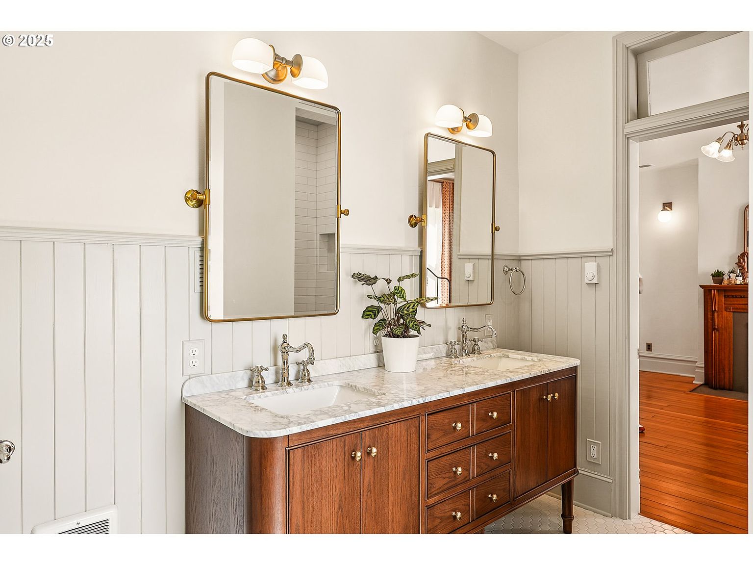 This is a well-lit primary bathroom featuring a double vanity with a marble countertop and dark wood cabinetry. Two gold-framed mirrors hang above the sinks, each illuminated by a double sconce. The walls are partially paneled, and the flooring appears to be hexagonal tiles, creating a classic and elegant aesthetic.