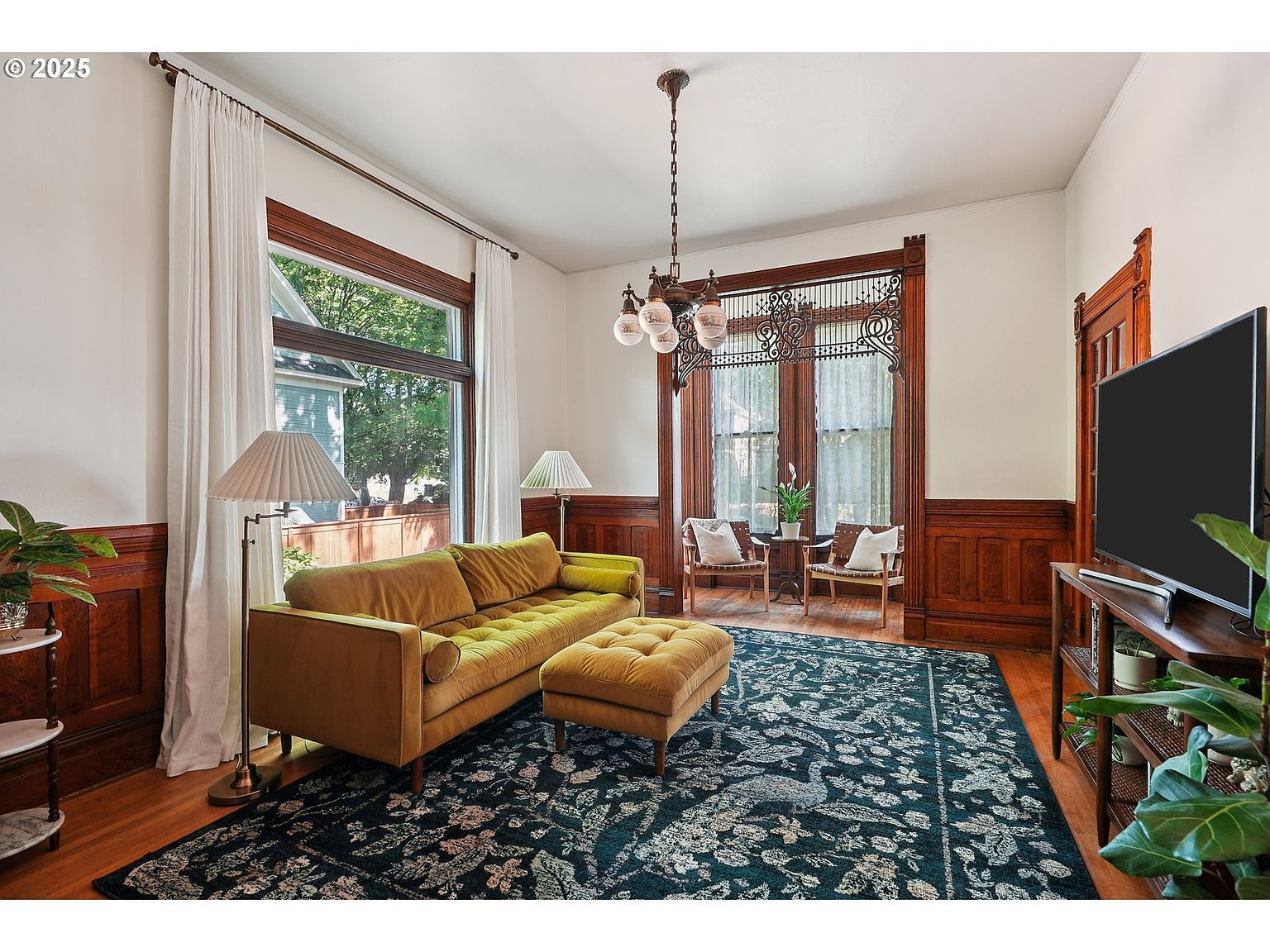 This is a beautifully staged living room featuring a mustard-colored sofa and ottoman set on a dark floral rug. The room is accented with dark wood trim and paneling, complemented by white walls and sheer curtains. Natural light floods the space, highlighting the hardwood floors and creating a warm, inviting atmosphere.