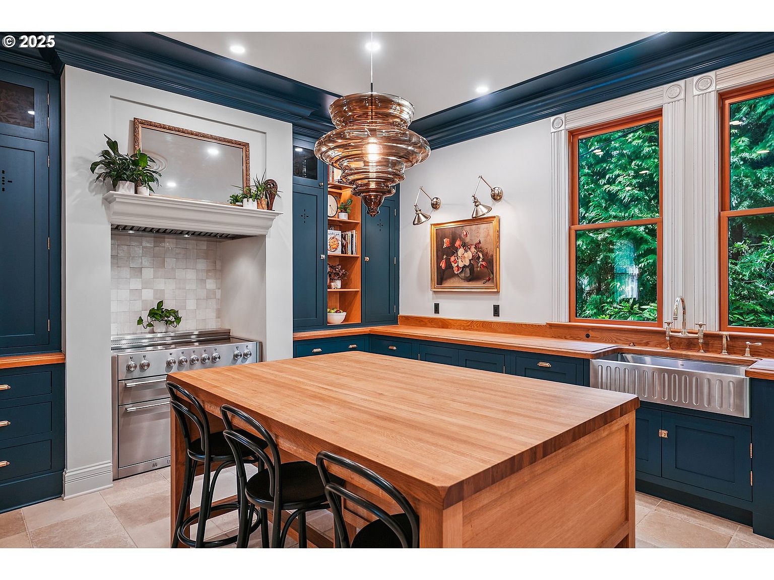 This is a well-appointed kitchen featuring custom blue cabinetry with wood countertops and a large wooden island with seating. Stainless steel appliances and a decorative range hood add to the upscale feel, while a large window provides natural light and a view of the outdoors. The kitchen is illuminated by a unique pendant light fixture, and the overall design is both elegant and functional.
