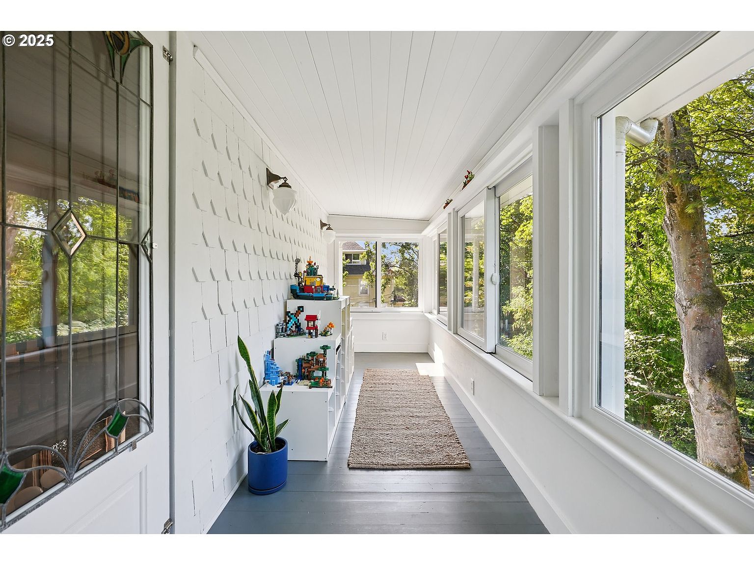 This bright sunroom features white walls and ceiling, with a series of windows offering views of the surrounding greenery. A neutral-toned rug runs along the floor, and a white shelving unit displays colorful toys and a potted plant. The room has a cozy and inviting atmosphere, perfect for relaxation or play.